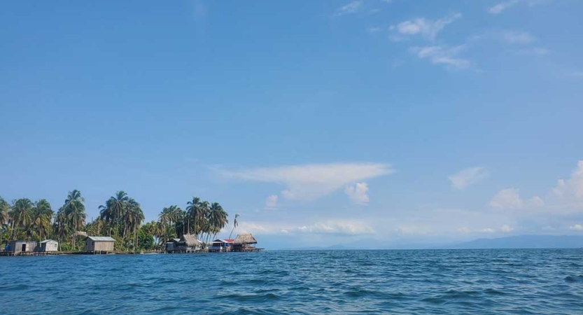 Blue ocean water extends far out to the horizon, beyond a small piece of land dotted with trees and structures 